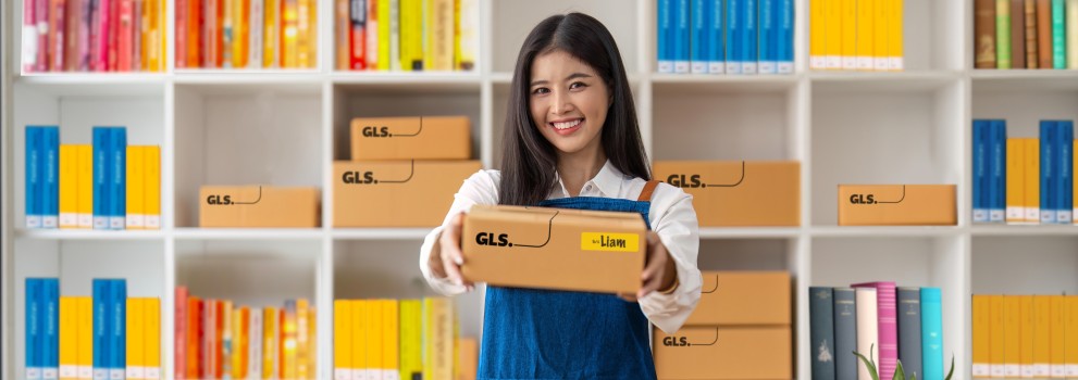 Woman handing out a GLS parcel. She stands at a desk in front of shelves field with colourful books. Woman handing out a GLS parcel. She stands at a desk in front of shelves field with colourful books.