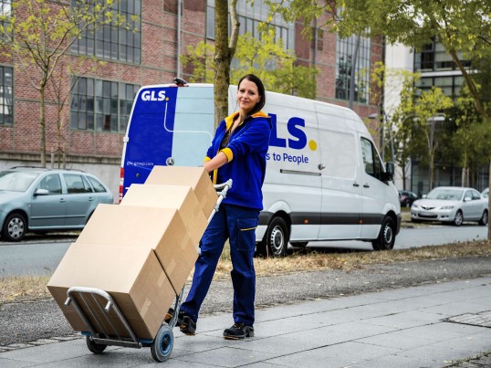 Livreuse GLS transportant plusieurs colis sur un diable, avec un fourgon de livraison GLS en arrière-plan.