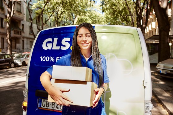 Une livreuse GLS sourit face à l'objectif en tenant deux colis devant une camionnette de livraison GLS stationnée dans une rue arborée. Des immeubles résidentiels et des voitures sont visibles en arrière-plan.