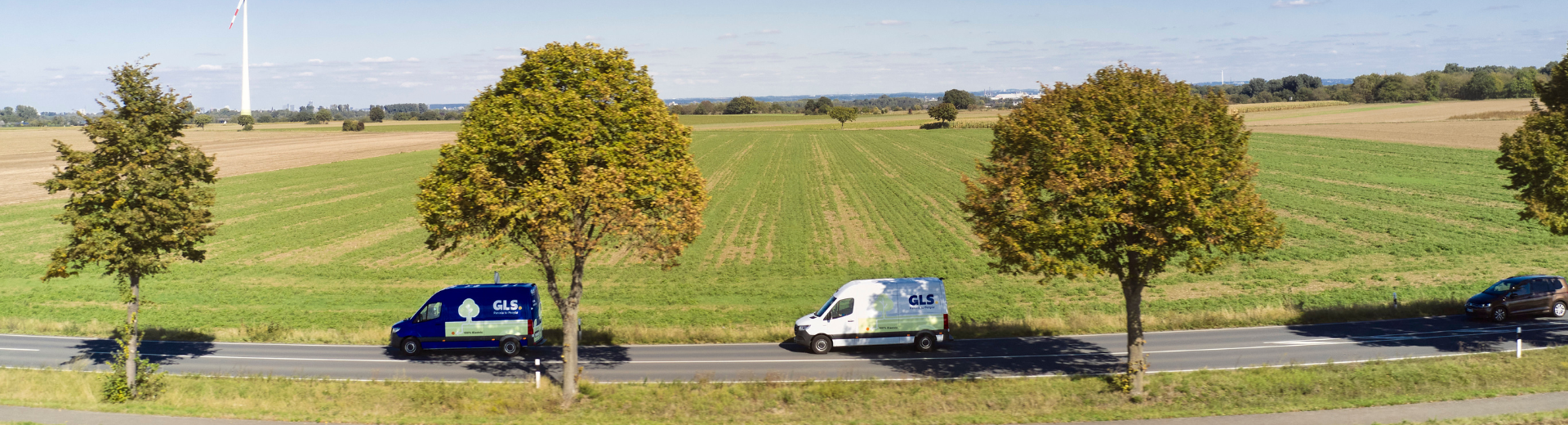 Deux camionnettes de livraison GLS sur une route de campagne, avec champs verts et éoliennes à l'horizon.