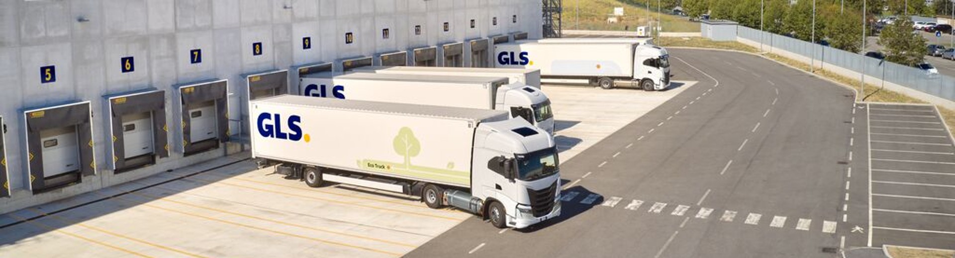 Een groot GLS-logistiek depot met laadperrons en geparkeerde vrachtwagens, gezien van boven, naast verharde wegen en grasvelden.