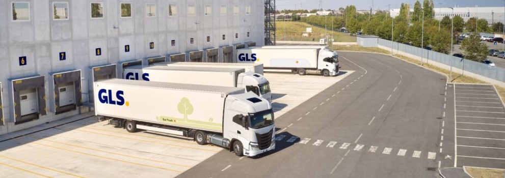 Een groot GLS-logistiek depot met laadperrons en geparkeerde vrachtwagens, gezien van boven, naast verharde wegen en grasvelden.