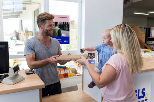 A woman holding her daughter hands a package to a man working at a parcel shop.