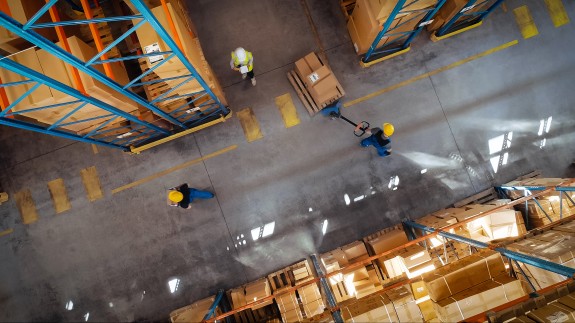 Overhead view of three warehouse workers performing separate tasks among aisles of stacked boxes.