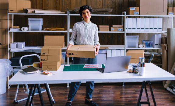 A woman standing at a table in a small warehouse, holding a cardboard shipping box. The workspace is filled with shelves of packing materials, stacked boxes, and shipping supplies.